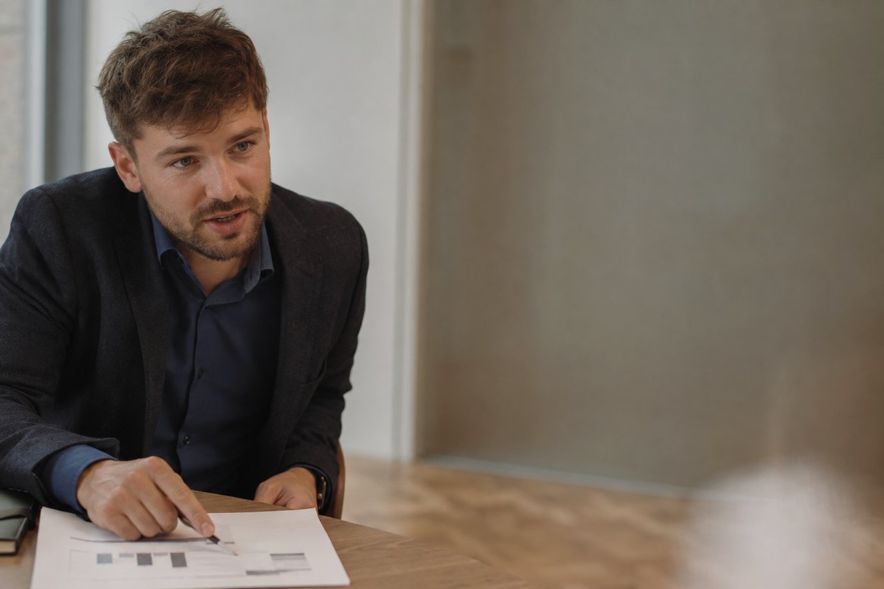 A businessman in a dark blazer and blue shirt sitting at a wooden table and pointing with his finger at a chart on a paper document during a conversation with a person seated opposite him