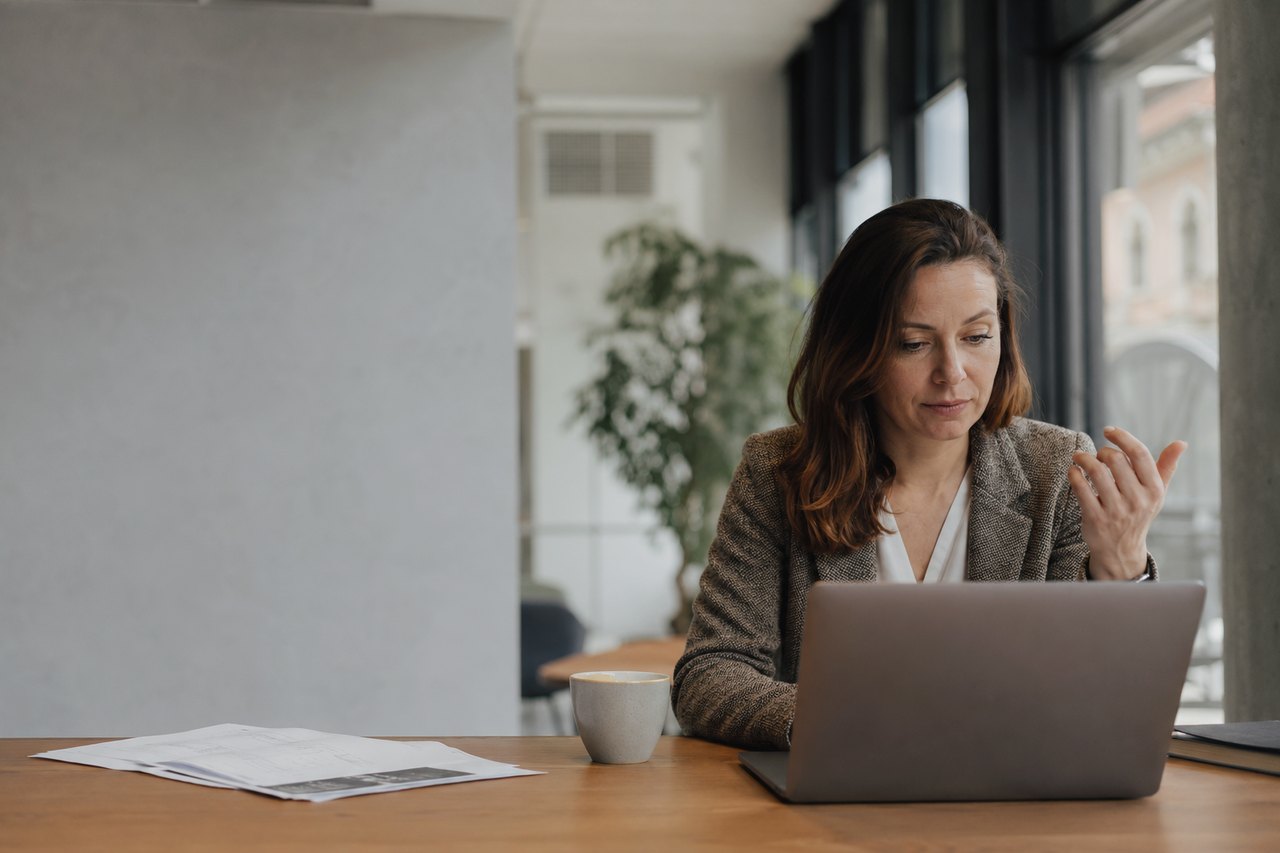 A businesswoman in a grey blazer sits at a wooden table with a laptop, a coffee cup and papers, in a bright office with a plant and large windows