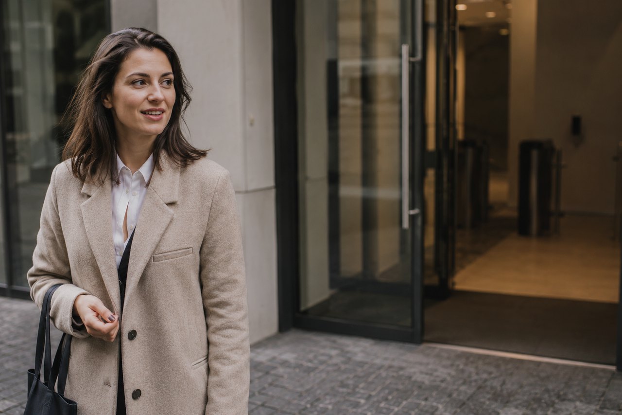 A businesswoman in a beige coat walks along a cobbled pavement in front of the entrance to a modern glass office building in central Prague; a lit foyer is visible through the glass doors.