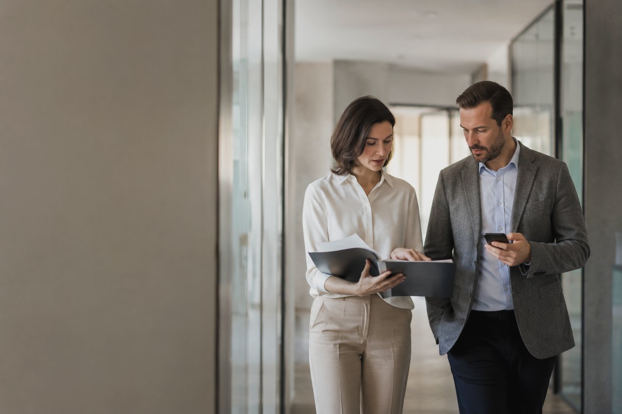 A businessman in a grey blazer and a businesswoman in a white blouse walking down a bright glass-walled office corridor, looking through documents in a folder together