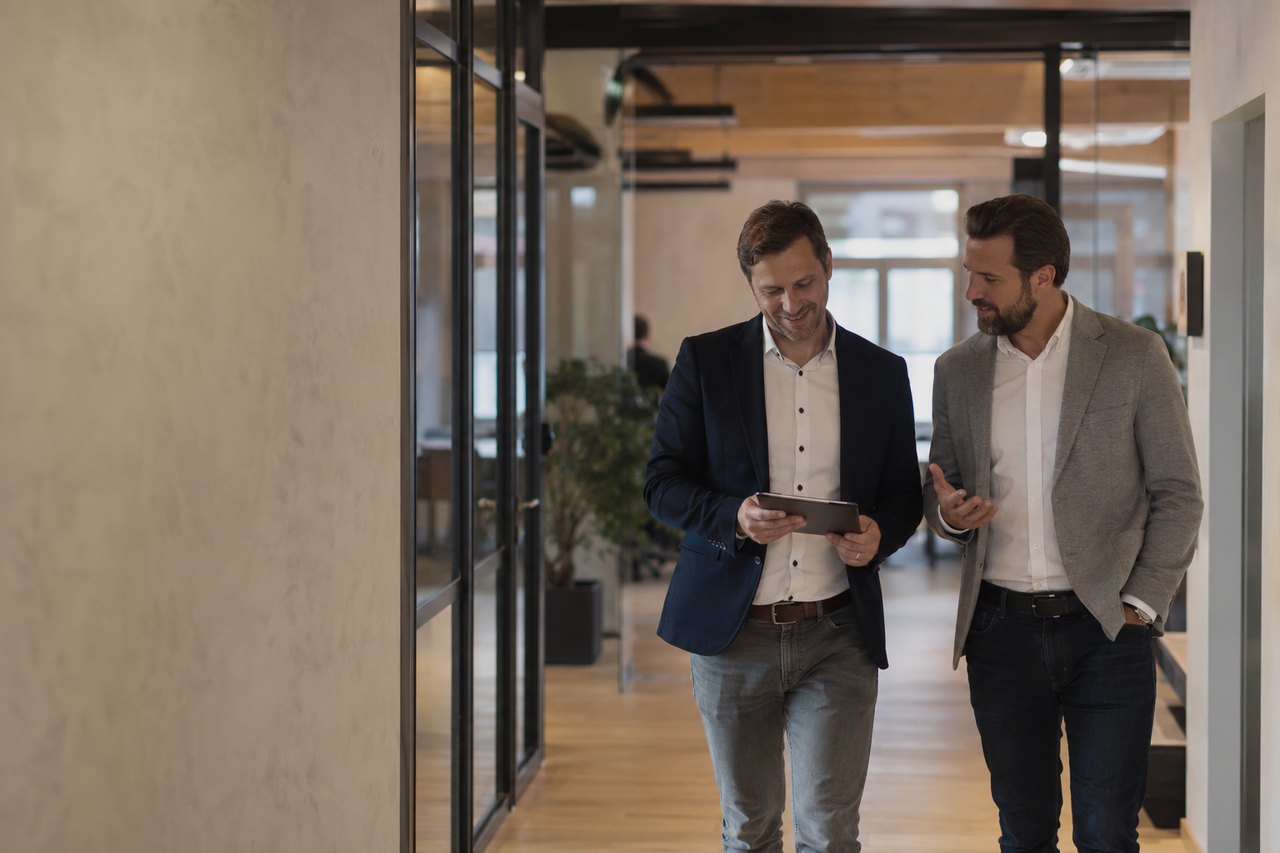 Two businessmen in blazers and white shirts walk down a light-filled corridor of a modern Prague office, talking over a tablet; glass-walled offices with a wooden frame on the left, a wooden floor and green plants on the right