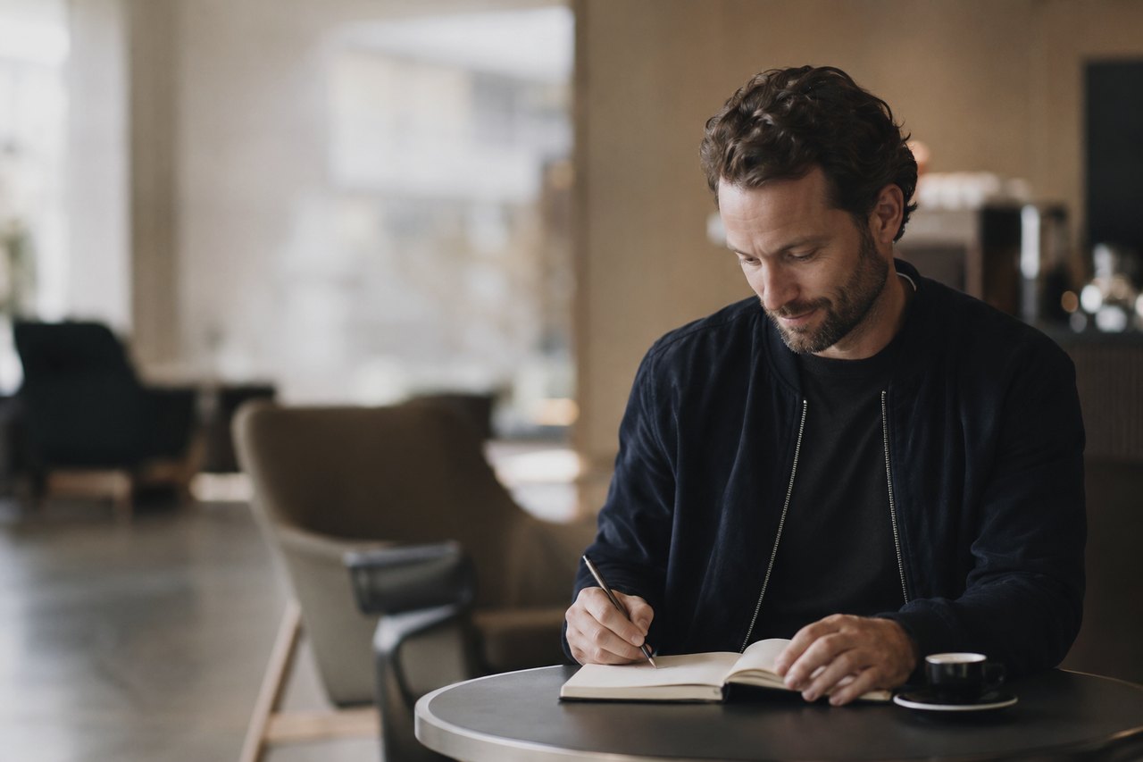 A man with a short dark beard in a navy jacket sits at a round table in a bright café, writing notes with a pen into an open notebook in concentration; a small cup of espresso stands beside him, with armchairs and a large window blurred in the background.