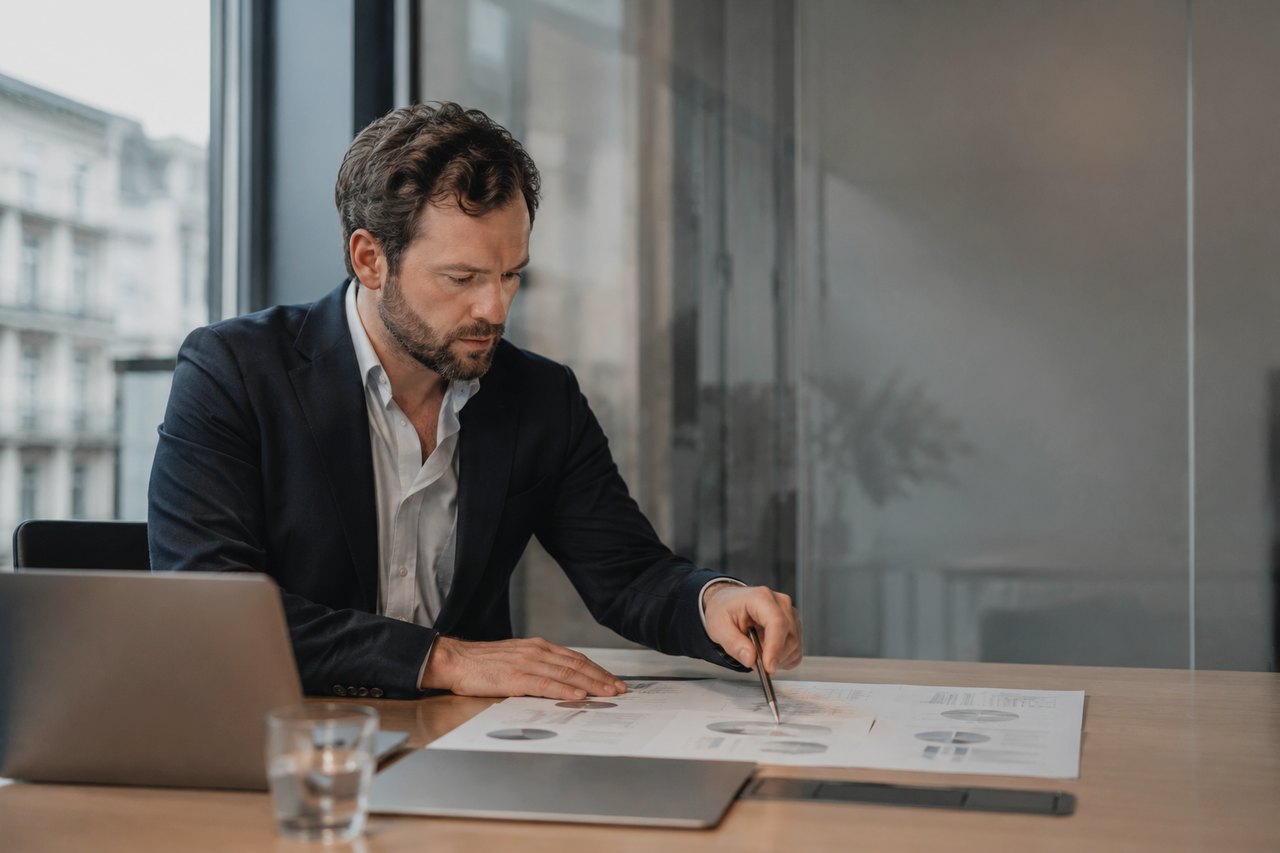 A businessman in a dark blazer sits at a table in a bright office, going through printed documents with charts using a pen, with an open laptop and a glass of water beside him