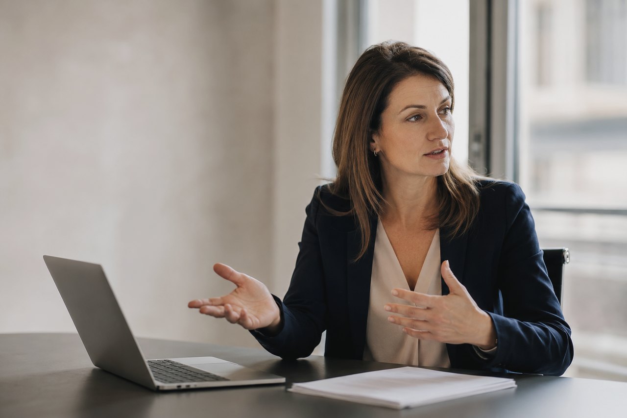 Businesswoman in a dark blazer sitting at a desk with a laptop and documents by an office window, explaining something during a conversation