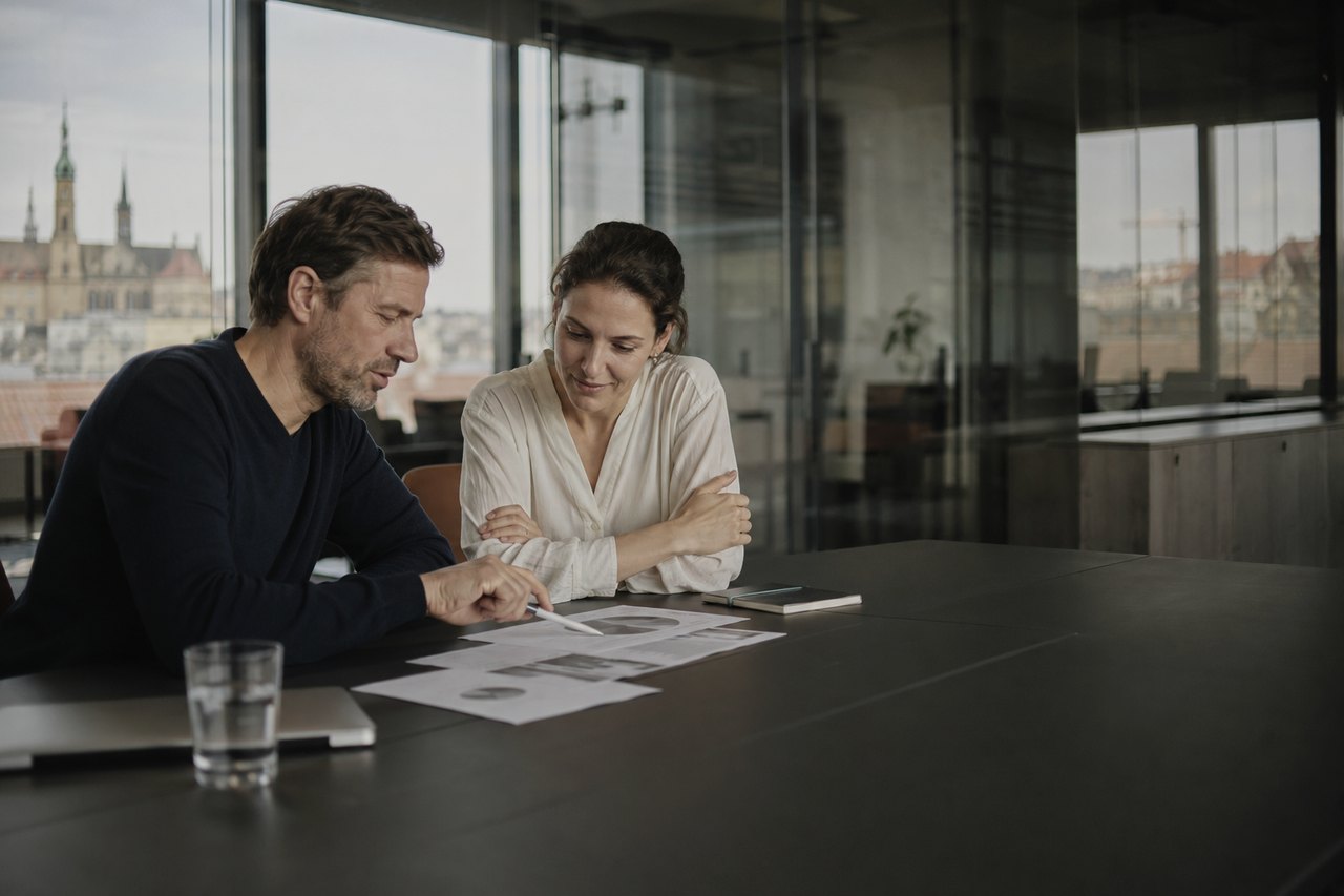 A man in a dark navy sweater and a woman in a white blouse sit at a dark table in a light-filled Prague office, going through printed documents together; a Prague skyline with towers and rooftops beyond the glass wall