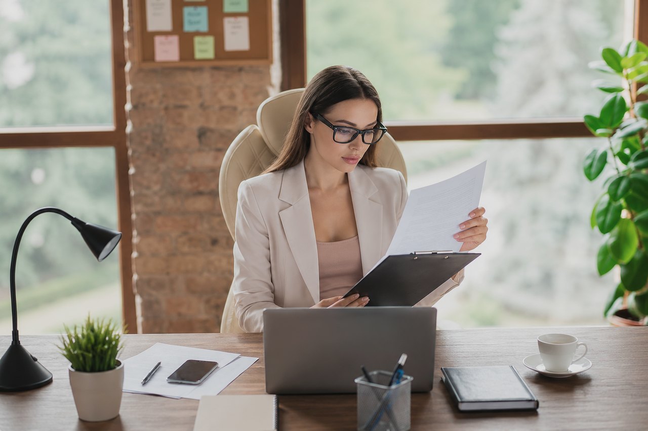 A businesswoman in glasses reading a document at her laptop in a modern office