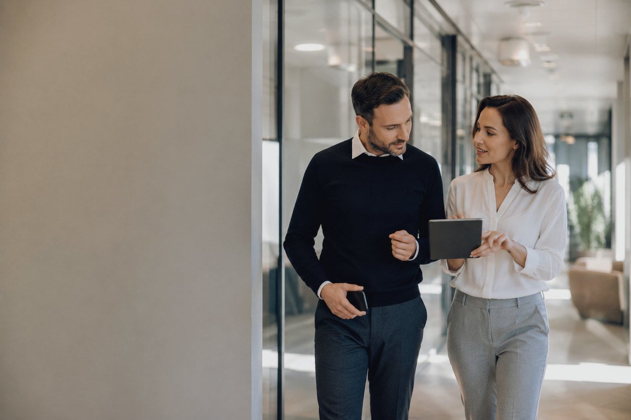 A businessman in a dark sweater and a businesswoman in a white blouse walking down a bright glass-walled office corridor, looking at a tablet together