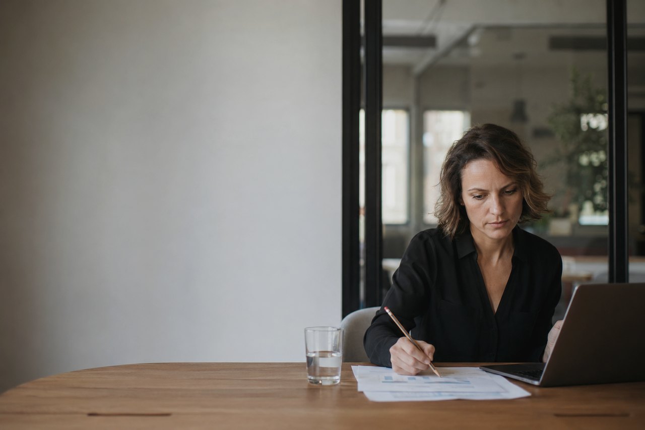 A woman in a black shirt sits at a wooden table, going through printed charts and tables with a pencil, an open laptop and a glass of water beside her; a glass wall and a bright modern office in the background.