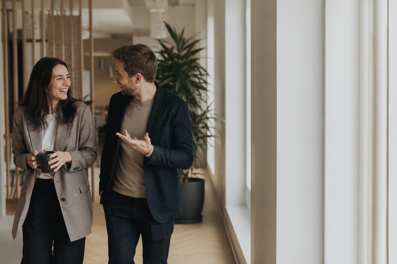 A businesswoman in a beige blazer holding a coffee mug and a businessman in a dark navy blazer walking and chatting with a smile in a bright corridor of a modern office with a plant and white pillars