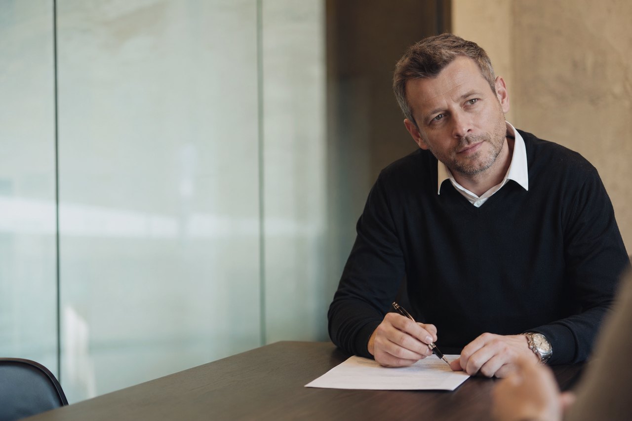 A businessman in a dark sweater and white shirt sitting at a dark wooden table and signing a paper document with a pen during a business meeting