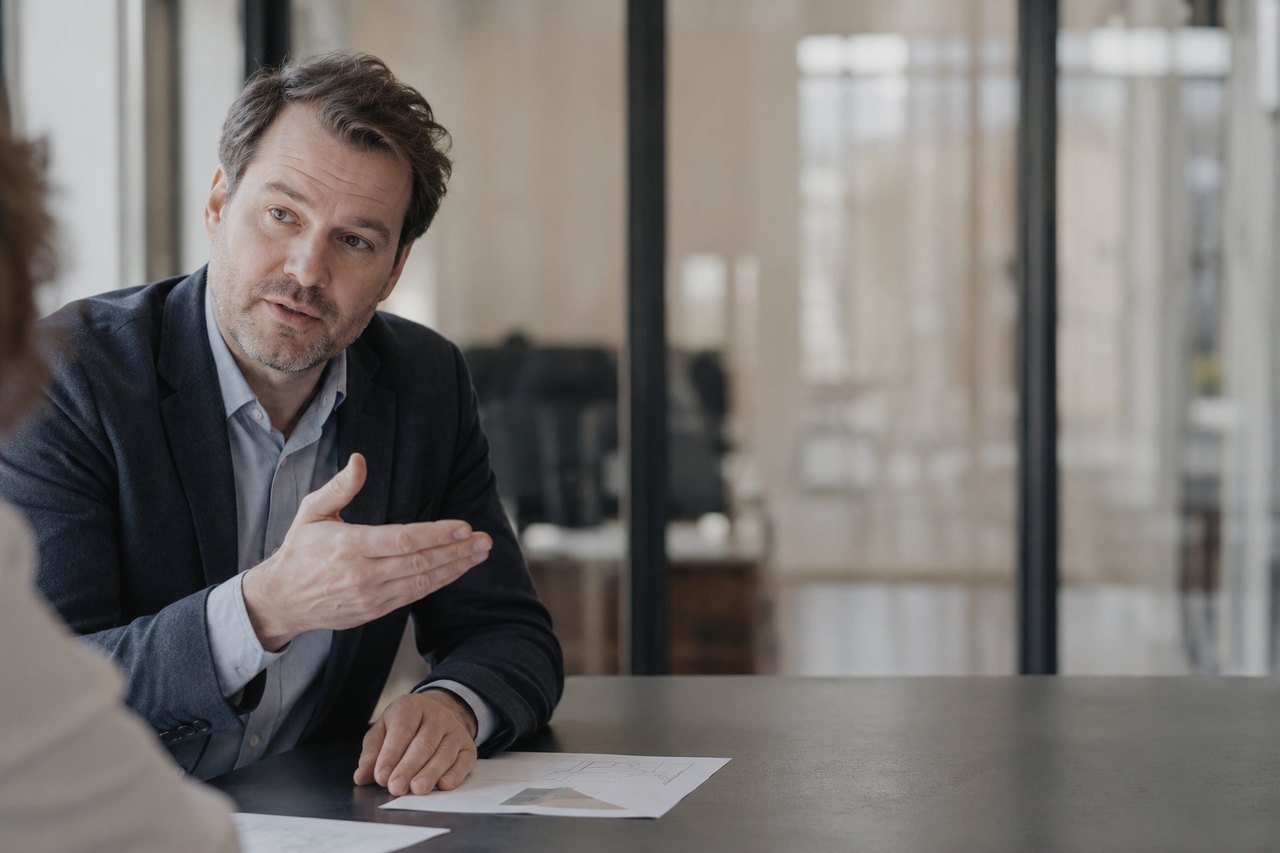 A businessman in a dark navy blazer at a table gesturing with his hand and explaining something to a partner sitting opposite, with documents and a sketch lying on the table in front of them, a bright glass-walled office in the background