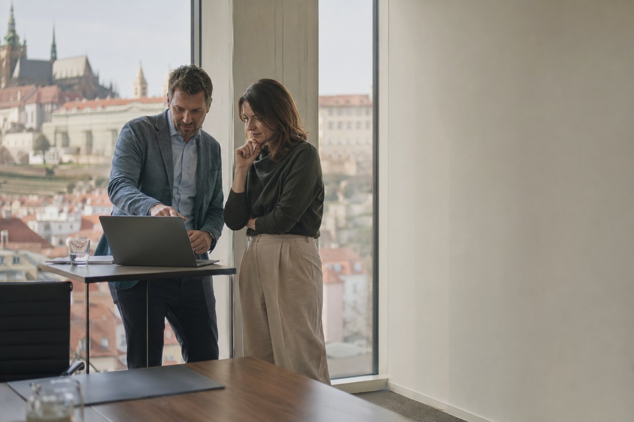 A businessman in a grey blazer and a businesswoman in a dark green sweater stand by the large glass windows of a Prague office, looking together at a laptop resting on a high table; beyond the window, in daylight, stretches the Prague skyline with Prague Castle and St. Vitus Cathedral.