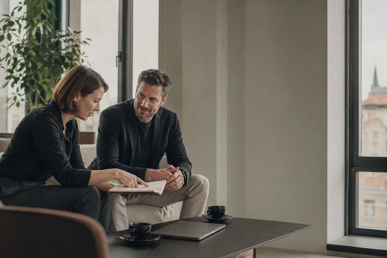 A businesswoman and a businessman in dark clothing sit at a small table reviewing notes in a notebook together, three coffee cups and a laptop on the table, Prague rooftops and spires through the window