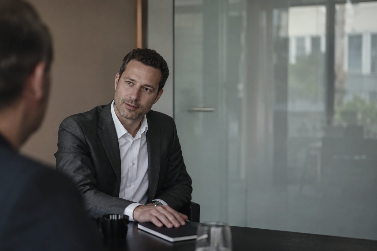 A businessman in a dark grey blazer and white shirt sitting at a dark table in a bright office with a glass partition, in conversation with another man seated opposite him