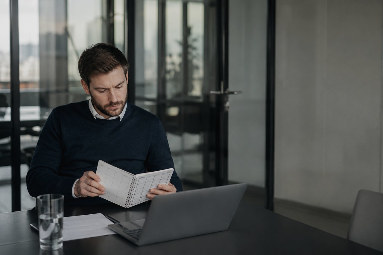 A businessman in a dark sweater sits at a table in a glass-walled office, looking through an open spiral planner, with a laptop and a glass of water in front of him