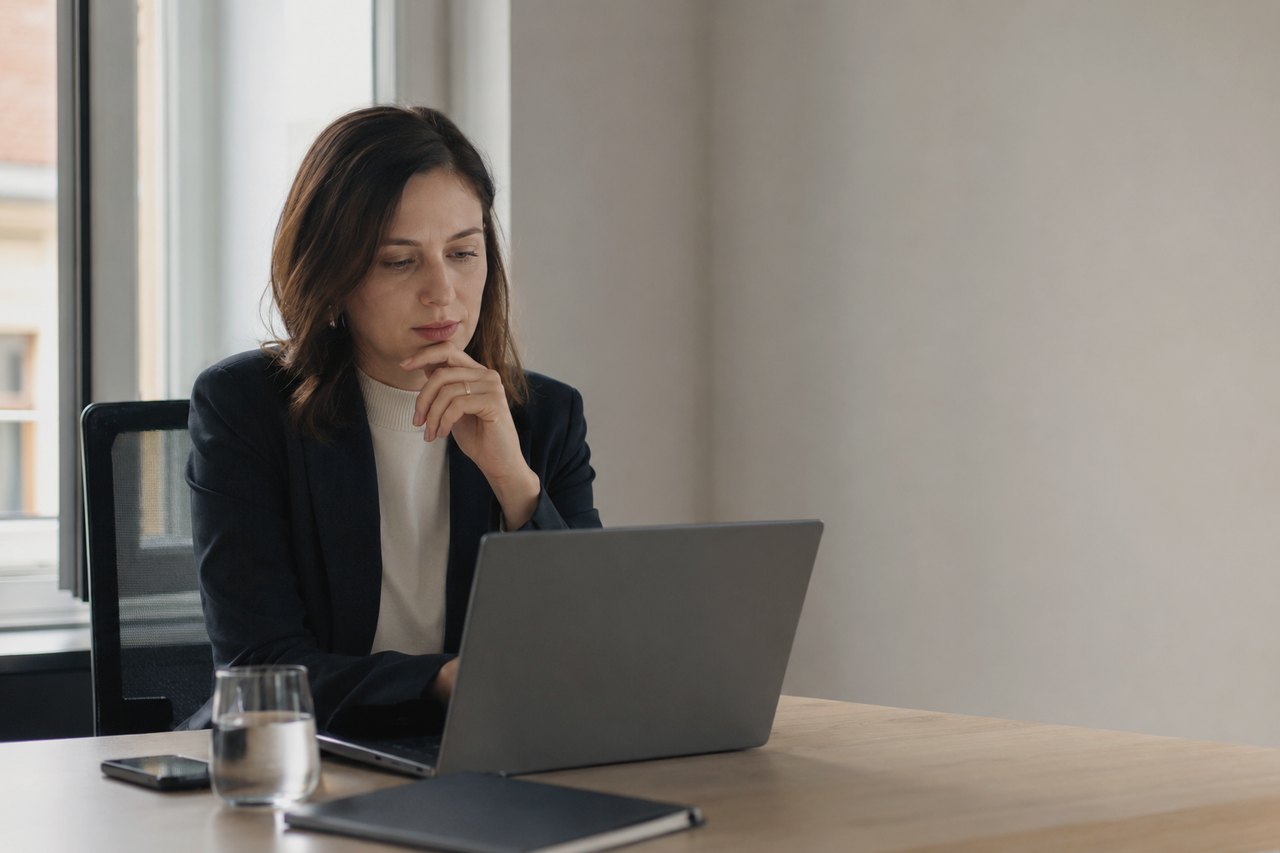 A woman in a dark blazer sits at a wooden table by a window, looking thoughtfully at a laptop with her hand resting on her chin; a glass of water, a phone and a notebook beside her in a bright office.