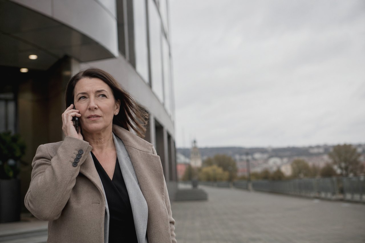 A businesswoman in a beige coat and grey scarf talks on the phone outside a modern Prague office building; behind her, under an overcast autumn sky, the panorama of Prague's rooftops with a church tower stretches out.