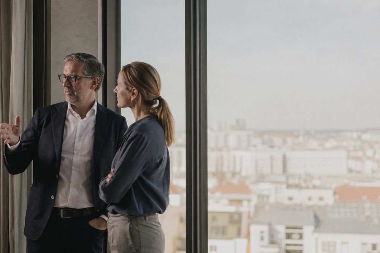 A grey-haired man in glasses and a dark blazer with a white shirt gestures in conversation with a woman with tied-back hair in a dark blouse; they stand by the glass wall of a high-rise office with a city skyline beyond the window