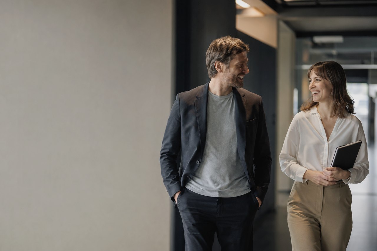 A smiling businessman in a dark navy blazer over a grey t-shirt walks beside a smiling businesswoman in a white blouse and beige trousers holding a black folder, down a bright modern office corridor with a light grey wall on the left and glass-walled offices on the right