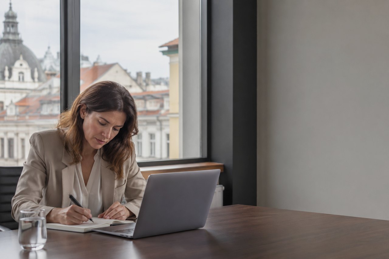 A businesswoman in a beige blazer sits at a wooden desk with a laptop and a notebook, taking notes, with a historic panorama of Prague's rooftops and a church dome in the large window