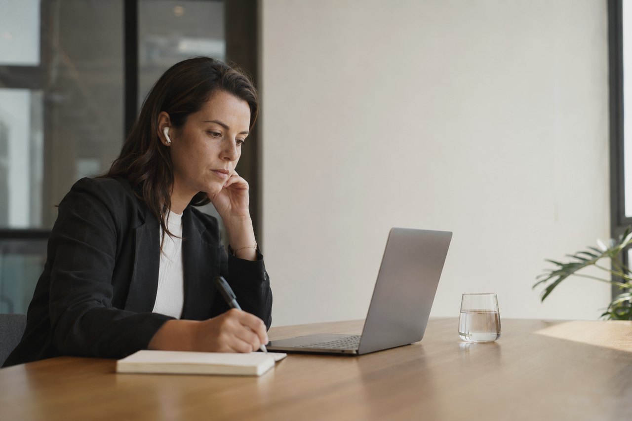 A woman in a blazer taking notes in a notebook beside a laptop in a bright office